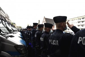 Umundurowani policjanci oddają honor poległemu na służbie policjantowi. Za nimi policyjne radiowozy.