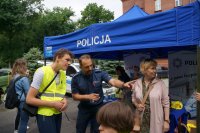 Dzieci w kamizelkach odblaskowych na tle stoiska raciborskiej policji rozmawiają z policjantem