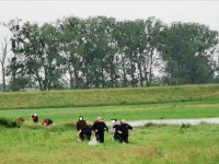 Raciborscy policjanci idą przez wysoką trawę z uratowaną załogą tratwy, a w oddali widać zacumowaną do brzegu tratwę