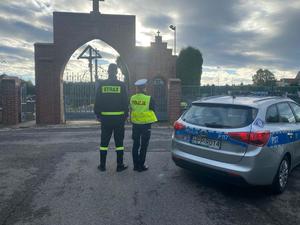 na zdjęciu policjantka radiowoz i strażak przy cmentarzu