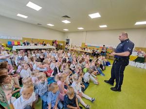 na zdjęciu przedszkolaki siedzą na dywanie i słuchają policjanta