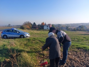 na zdjęciu seniorka podpierająca się miotła i objęciach syna. Policjant stoi z boku